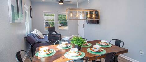 A dining area with a wooden table set for four, surrounded by chairs, a ceiling fan, and a mounted TV.