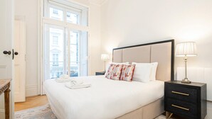 Un lit bien fait, recouvert d’une couette blanche, avec une paire de serviettes blanches et des coussins décoratifs. Il y a une table de chevet avec une lampe et un petit tiroir. La chambre est dotée d’une grande fenêtre donnant sur les bâtiments environnants.