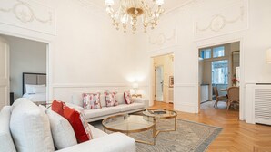 Un salon spacieux avec un canapé, une table basse et un lustre. On aperçoit à l’arrière-plan un coin repas avec une table et des chaises.