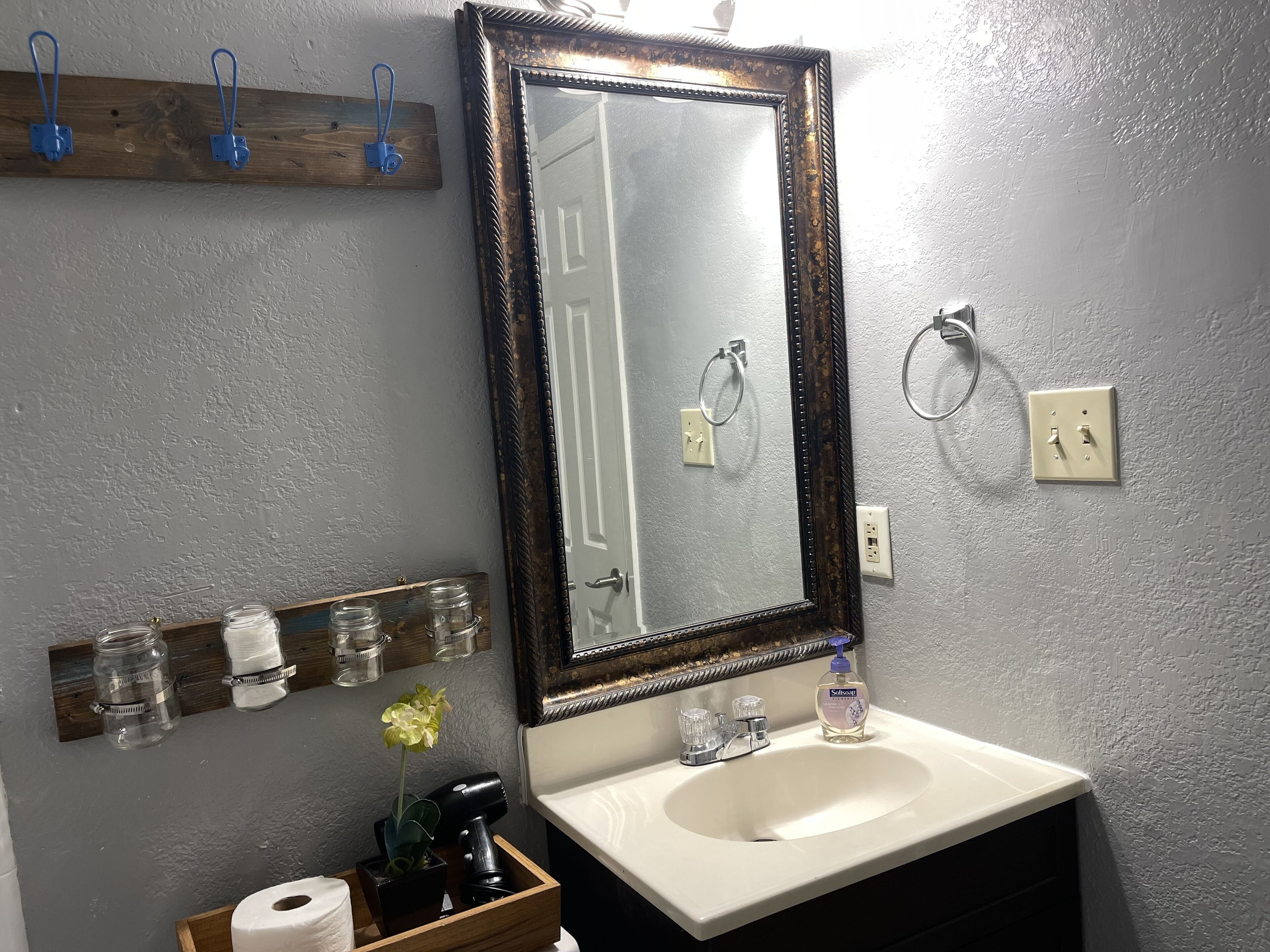 A bathroom with a white sink, a mirror with a dark frame, and a wooden shelf above the sink holding jars and a hairdryer.