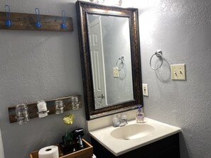 A bathroom with a white sink, a mirror with a dark frame, and a wooden shelf above the sink holding jars and a hairdryer. - TXST Cozy and centrally located (San Marcos)