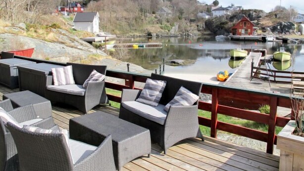 Une terrasse dotée de meubles en osier, donnant sur un lac paisible où naviguent des bateaux, et entourée de collines et de maisons.