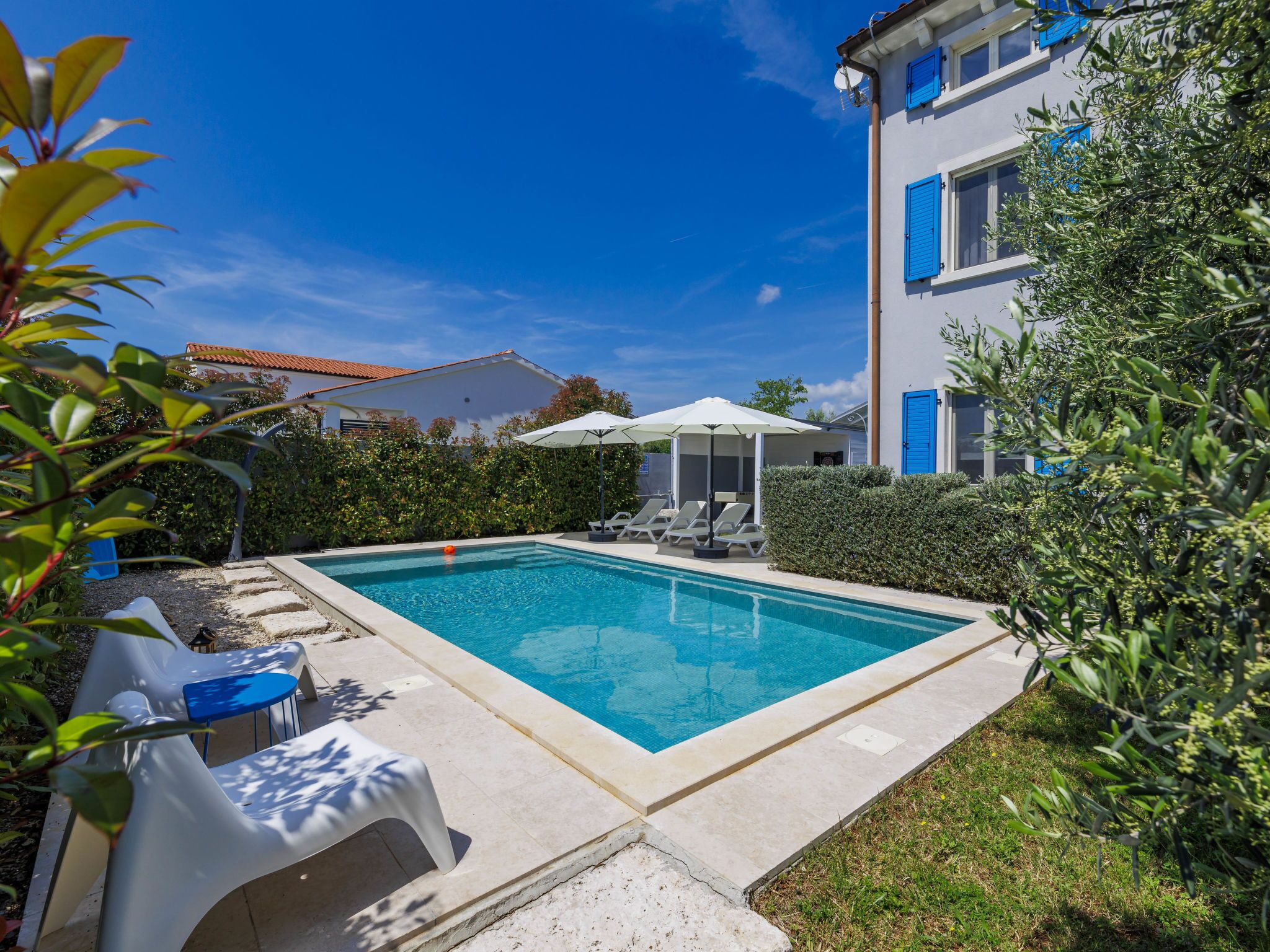 Une piscine rectangulaire dotée d’une plage blanche, entourée d’une haie et bordée d’un bâtiment aux volets bleus.
