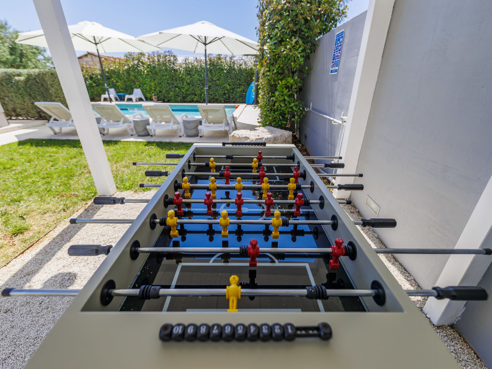 Une table de baby-foot installée à l’extérieur, avec une piscine et des chaises longues en arrière-plan.