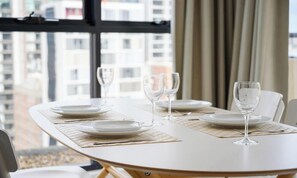 Une table à manger, dressée avec des assiettes et des verres à vin, est placée près d’une fenêtre offrant une vue sur la ville.