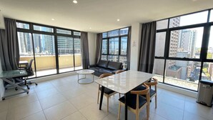 A modern living room with a sofa, a dining table, and a small coffee table. Large windows offer views of a cityscape. - MetaWiseBnB|Sydney CBD|Chinatown|Luxe 2Bed Penthouse Bella (Haymarket)
