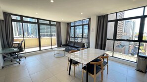 Ein modernes Wohnzimmer mit einer Couch, einem Esstisch und einem kleinen Couchtisch. Große Fenster bieten Blick auf die Stadt.