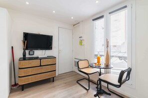 A room with a television, a rattan storage unit, a dining table with chairs, and a window with a view.