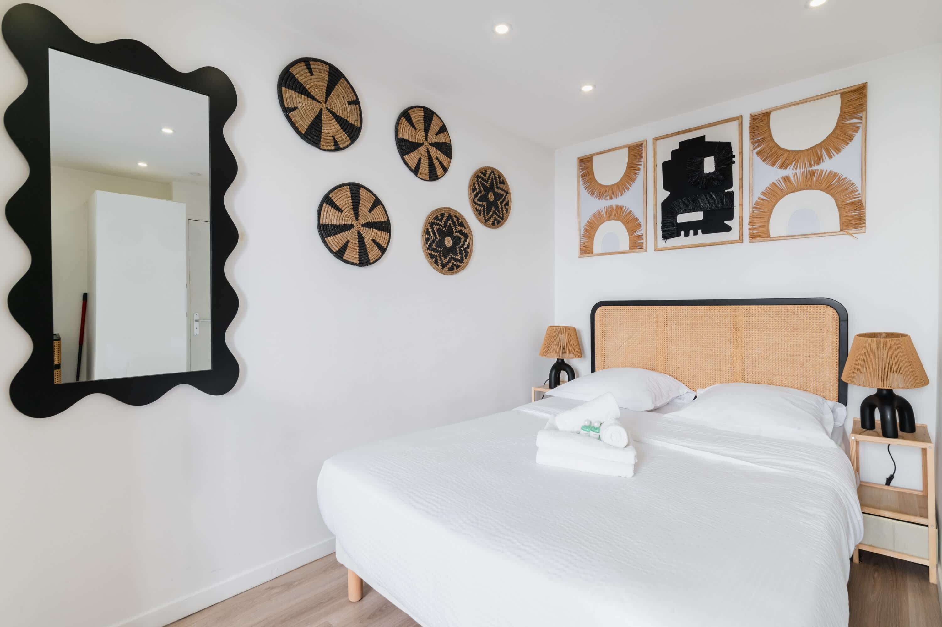 Una cama bien hecha con sábanas blancas, un espejo grande con marco negro y varias piezas de arte tejidas colgadas en la pared.