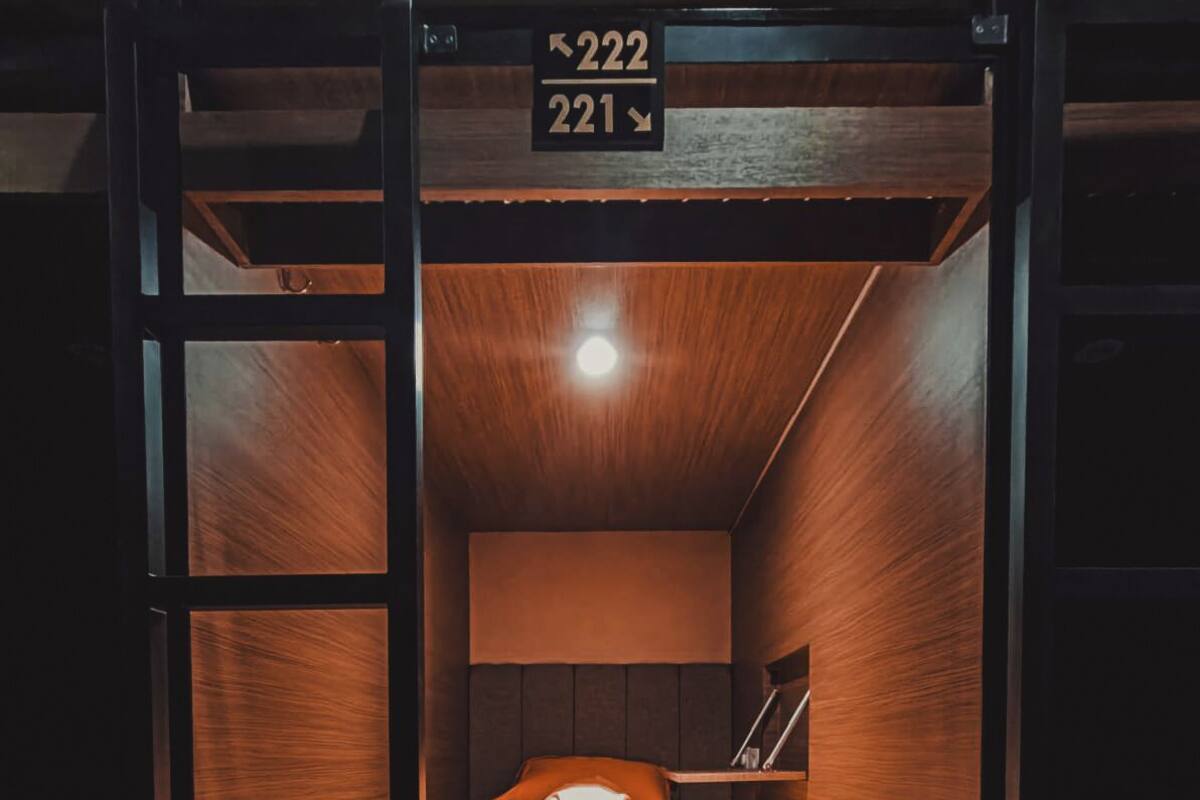 A bunk bed room with wooden walls and a slanted ceiling. There is a bed with a pillow and a blanket, and a small table with a lamp. The room has a number sign above the bed indicating room numbers 221 and 222.