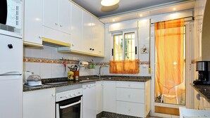 Een kleine keuken met witte kastjes, een inbouwoven, een koffiezetapparaat en een raam met decoratieve gordijnen.