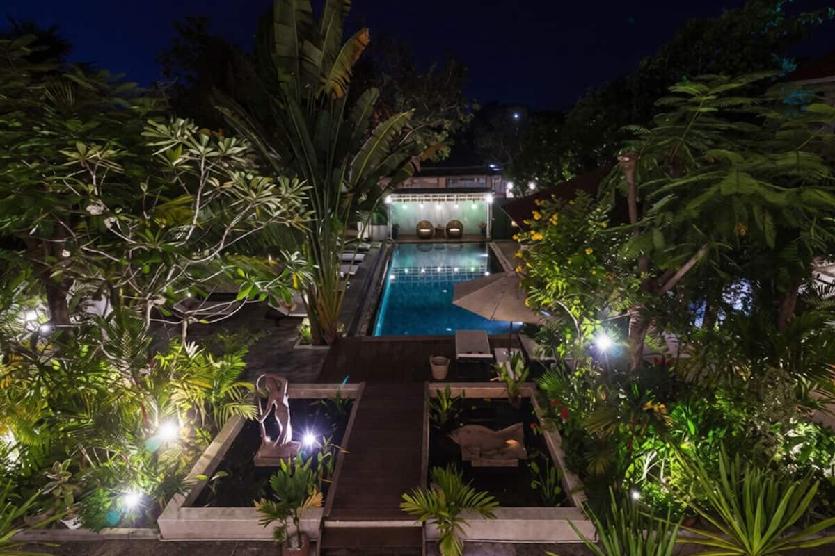 Vue nocturne d’un espace piscine avec une terrasse en bois, des chaises longues et un bâtiment en arrière-plan.