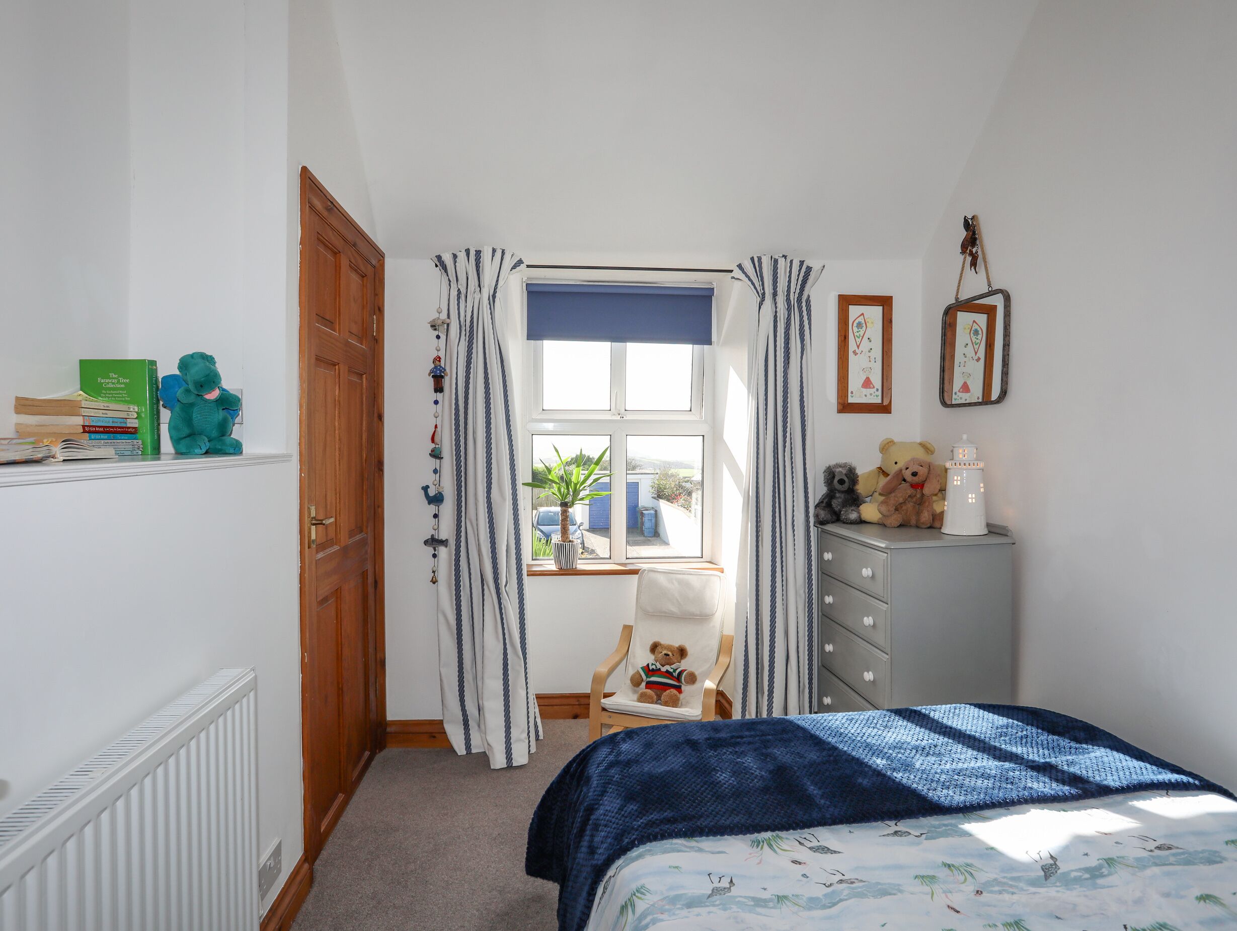 Une chambre à coucher comprenant un lit, une commode, une chaise, une fenêtre avec des rideaux et un radiateur.