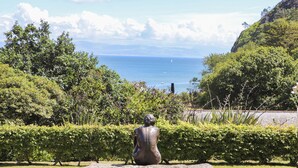 A sculpture of a seated figure in a garden with a view of the sea and mountains. - Bron Gadair (Pwllheli)