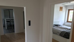 A room with a bed, a window, and a radiator. There is a door leading to another room with a kitchen area visible. - Large apartment with 2 bathrooms and 2 large parking spaces (Leichlingen (Rheinland))