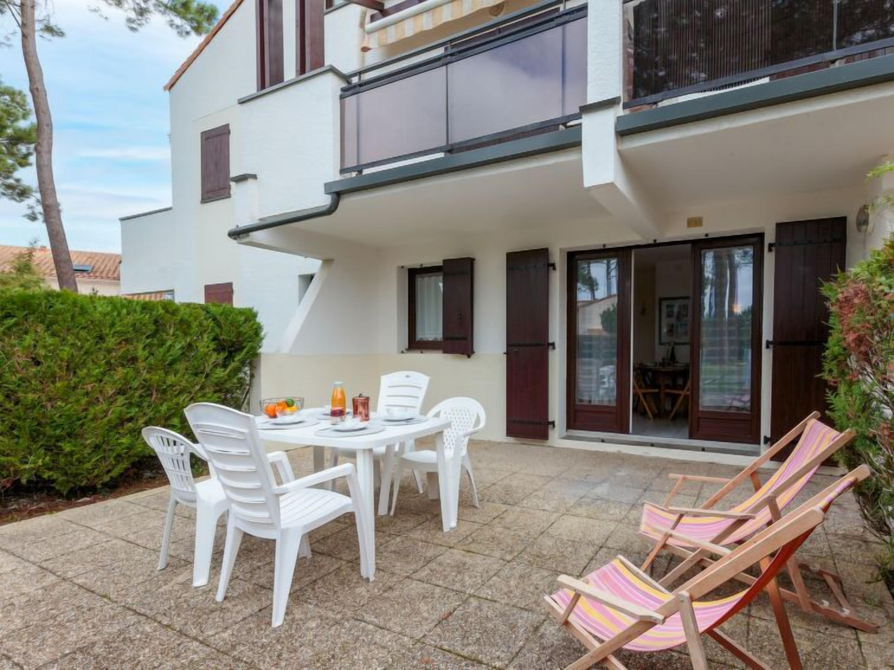 Un edificio moderno de dos pisos con un patio que incluye una mesa redonda y sillas de plástico blanco, una terraza de madera con sillas a rayas y un seto bien cuidado.