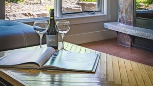 A room with a large window, a wooden table, two wine glasses, a bottle of wine, and a book. - Pooches Paradise - Pet Friendly Hot Tub & Pool (Canmore)