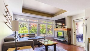 A living room with a sofa, a coffee table, a fireplace, and large windows. - Pooches Paradise - Pet Friendly Hot Tub & Pool (Canmore)