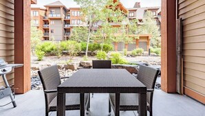 A covered patio with a table and chairs, overlooking a landscaped garden and a multi-story building with balconies. - Pooches Paradise - Pet Friendly Hot Tub & Pool (Canmore)