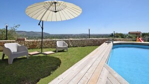 Ein Poolbereich mit Holzterrasse, zwei weißen Stühlen, einem großen Sonnenschirm und einer Steinmauer.