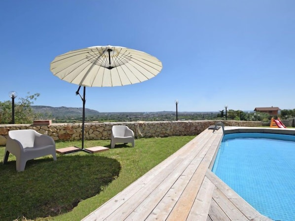 Un espace piscine doté d’une terrasse en bois, de deux chaises blanches, d’un grand parasol et d’un mur en pierre.