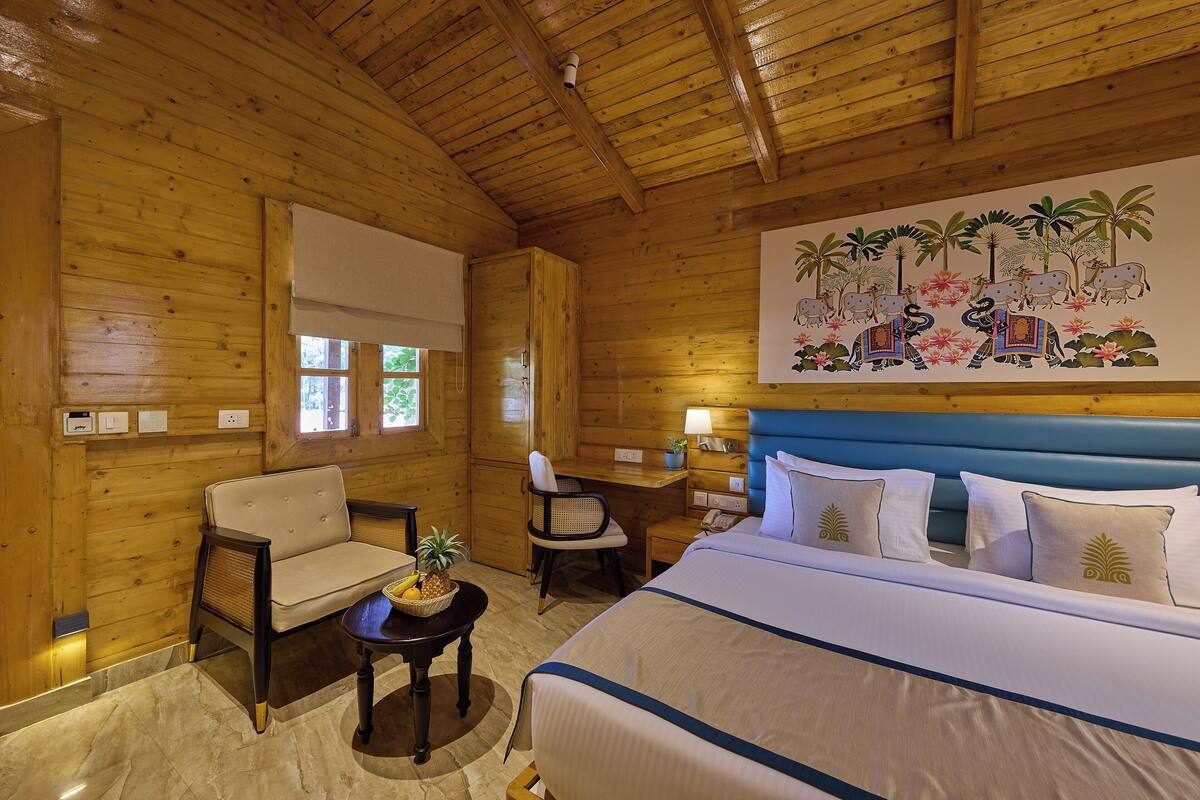 A wooden room with a bed, two chairs, a small table with fruit, and a painting of elephants and flowers on the wall.