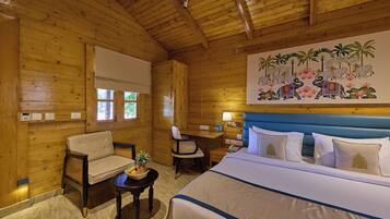 A wooden room with a bed, two chairs, a small table with fruit, and a painting of elephants and flowers on the wall.