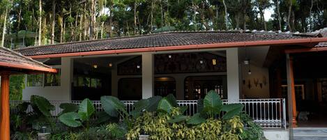 A covered outdoor area with a tiled floor, surrounded by large tropical plants and a stone pathway.