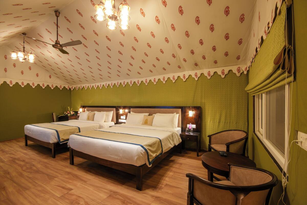 A hotel room with two beds, a small table, chairs, a ceiling fan, and a window with a telephone.