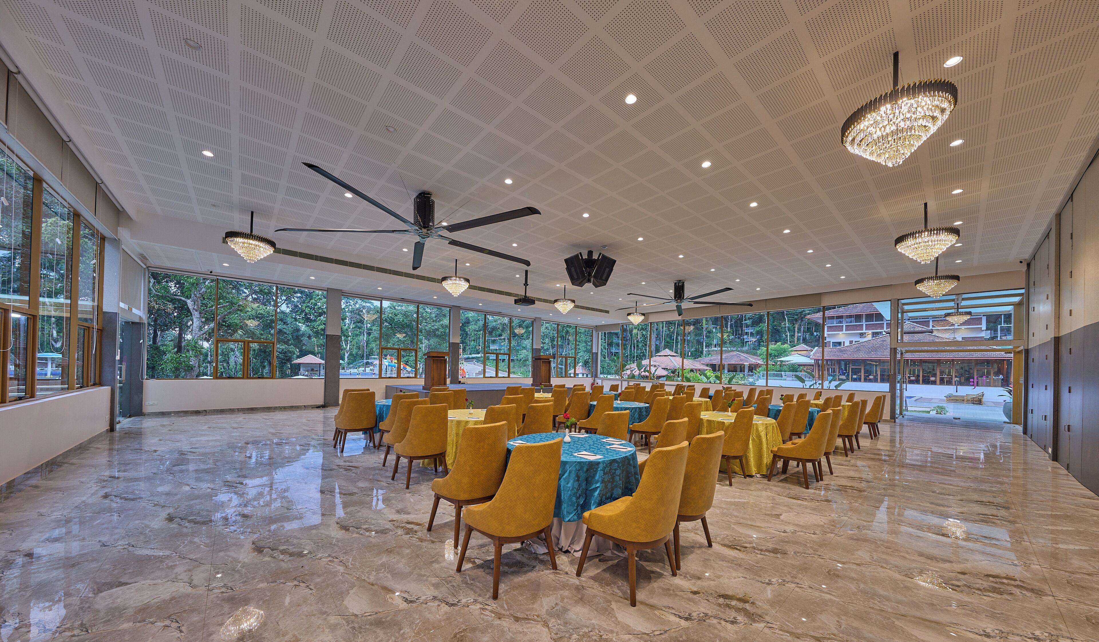 A spacious hall with large windows, yellow chairs, and tables, ceiling fans, and chandeliers.
