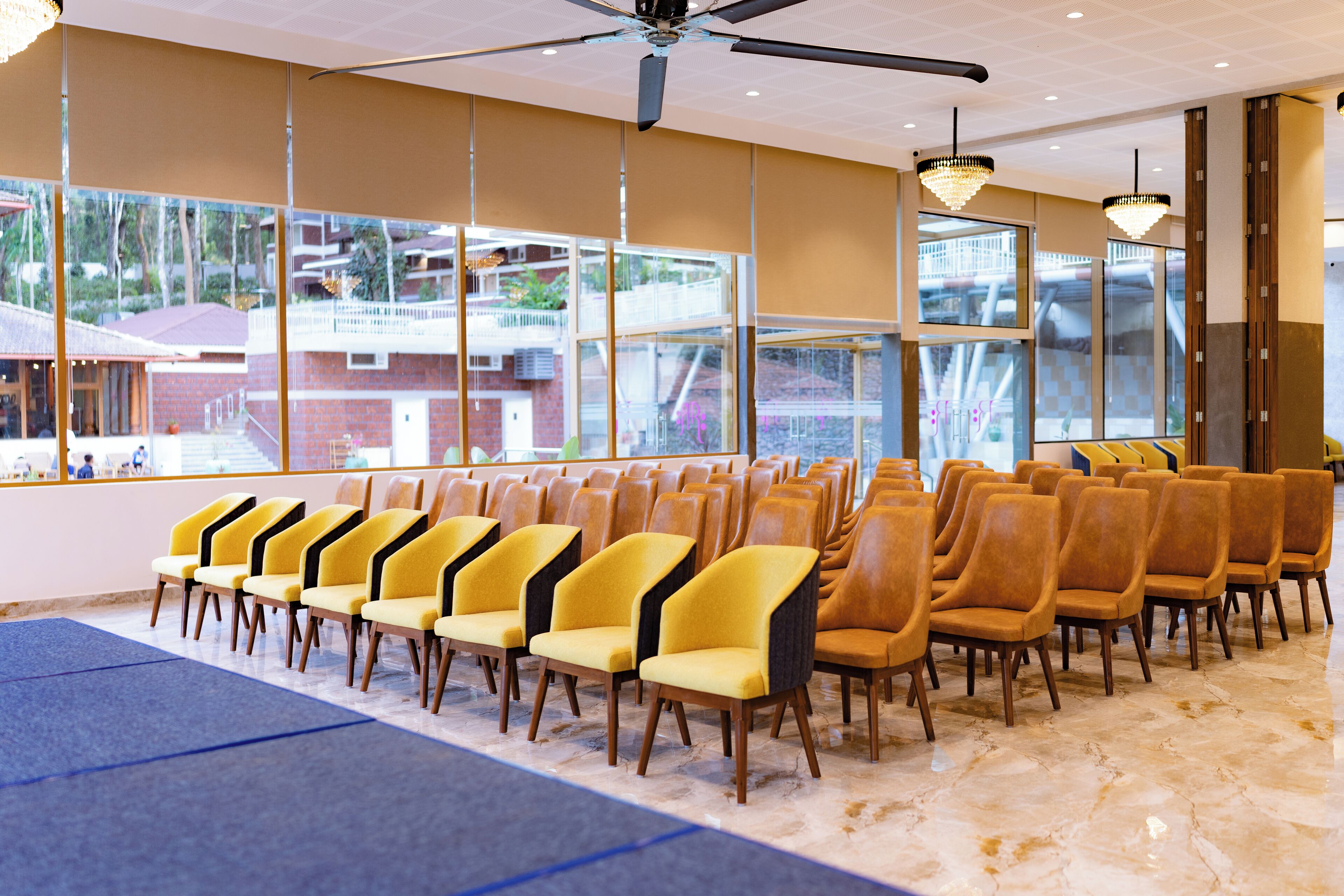 Un salón de conferencias con filas de sillas amarillas, ventanas amplias y un ventilador de techo.
