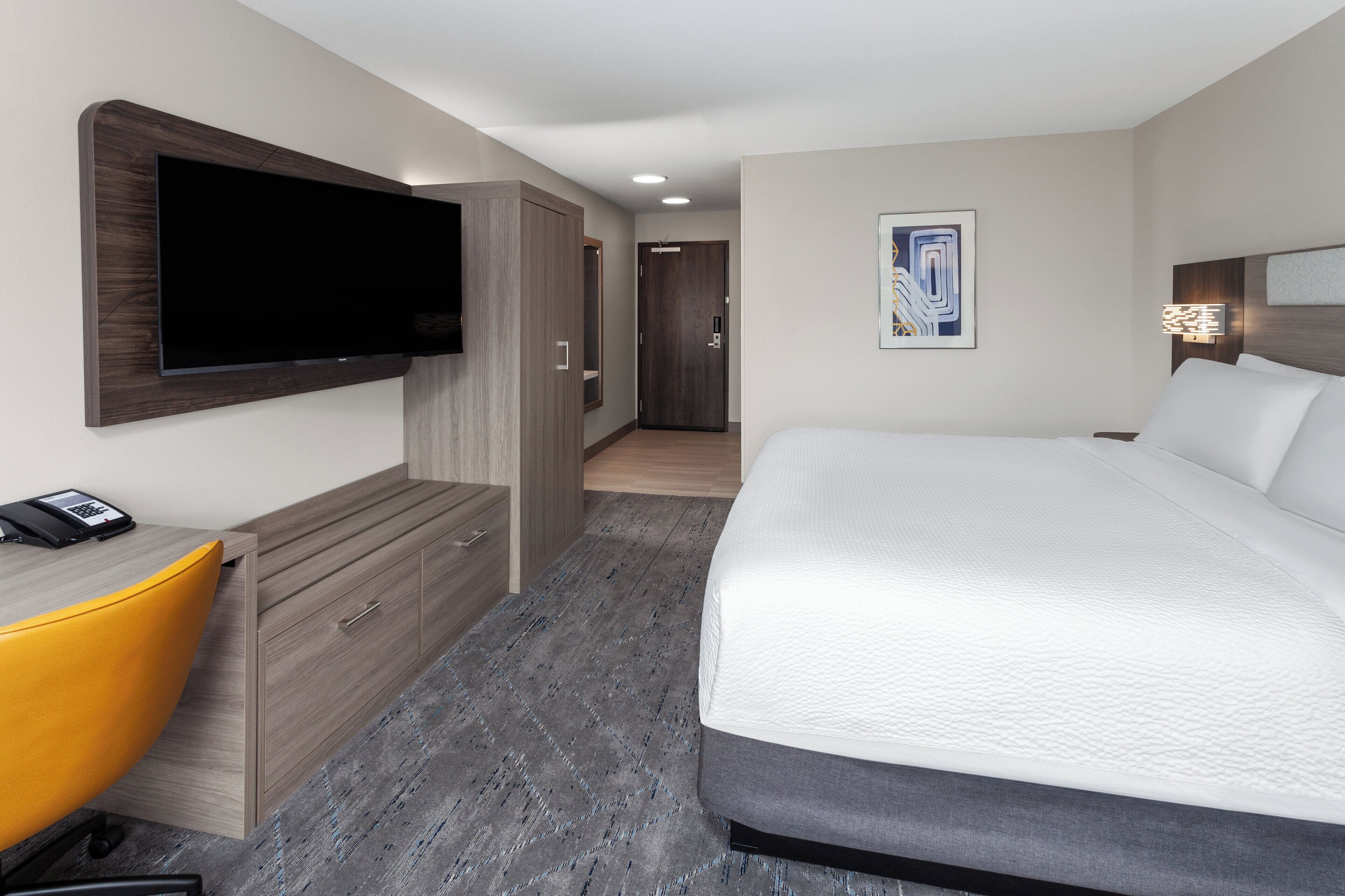 A hotel room with a bed, a desk with a chair, a television, and a door leading to another room.