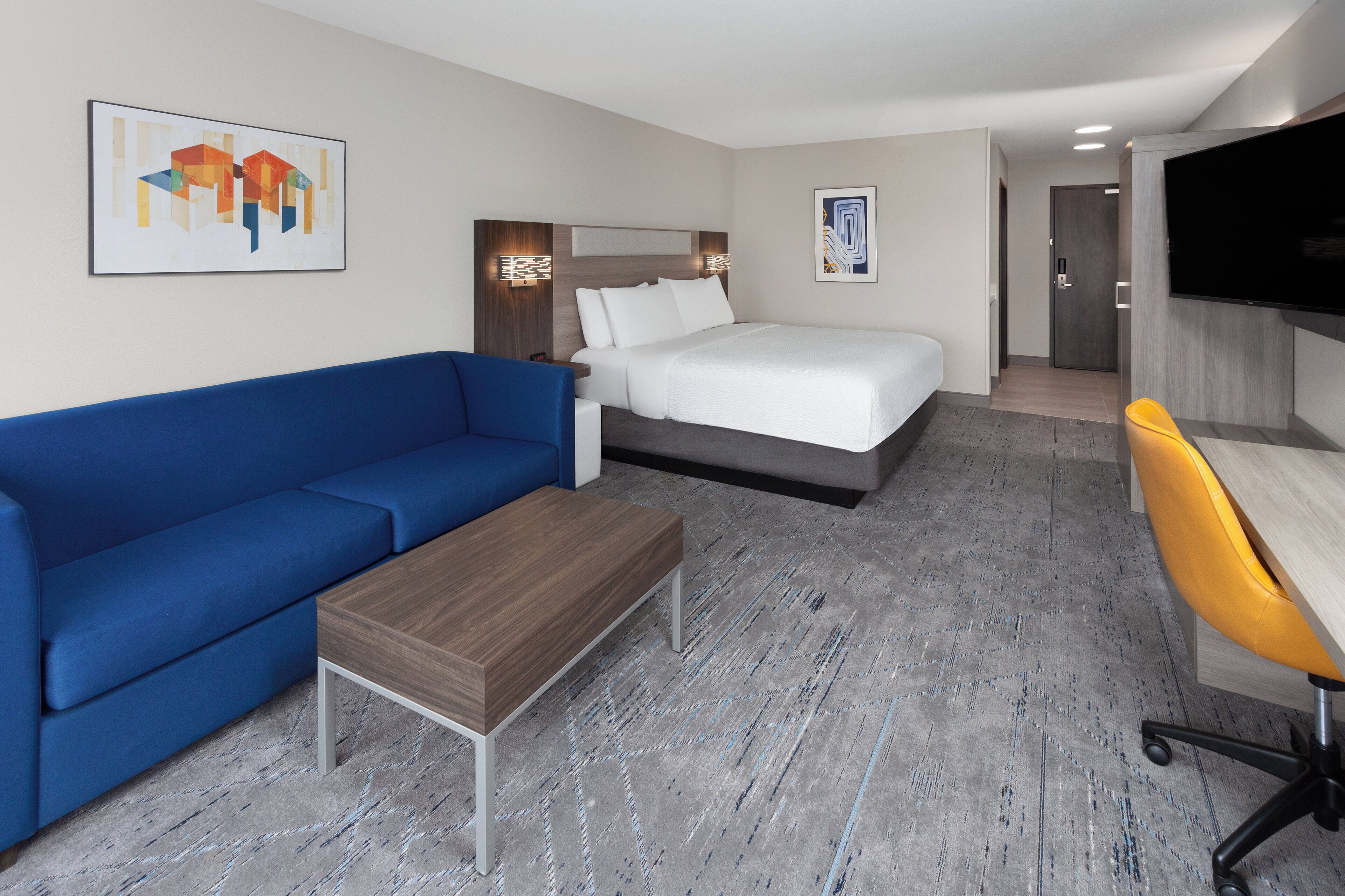 Une chambre d’hôtel avec un lit, un canapé bleu, une table basse en bois, un bureau avec une chaise jaune et un téléviseur fixé au mur.