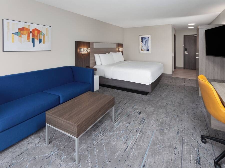 Une chambre d’hôtel avec un lit, un canapé bleu, une table basse en bois, un bureau avec une chaise jaune et un téléviseur fixé au mur.