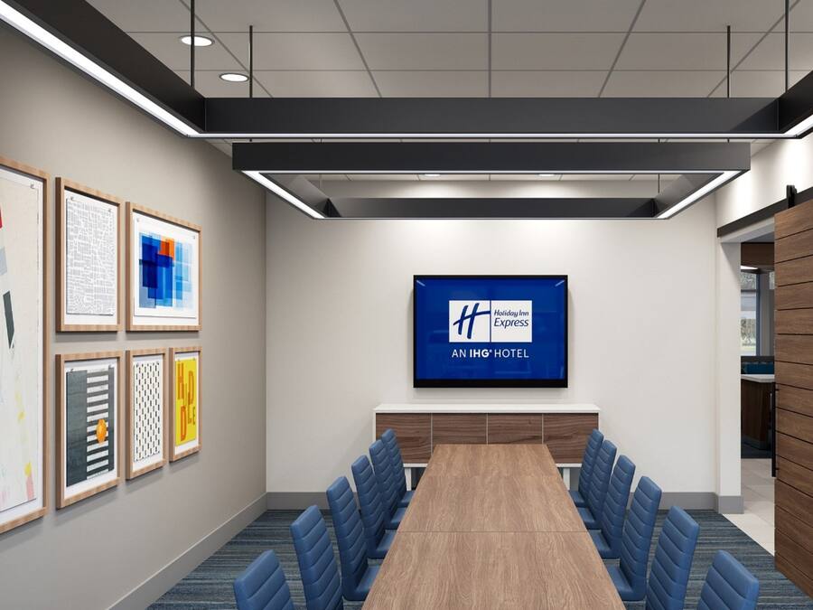Uma sala de conferências com uma mesa comprida, cadeiras azuis, obras de arte emolduradas nas paredes e uma tela grande exibindo o logotipo Holiday Inn Express.