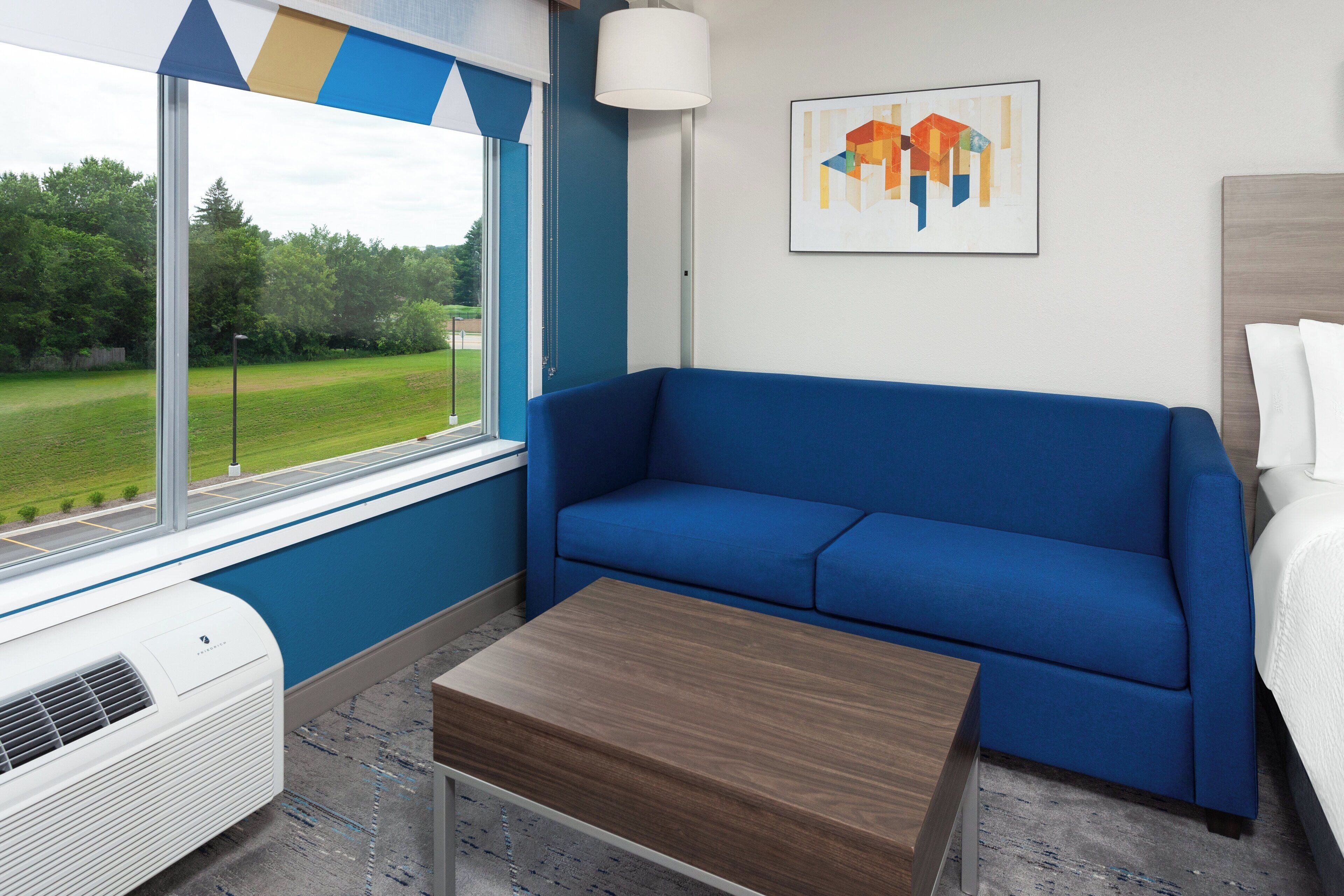 A room with a blue sofa, a wooden table, a window with a view of a green landscape, and a painting on the wall.