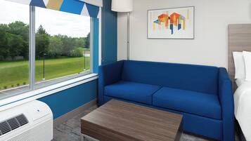 A room with a blue sofa, a wooden table, a window with a view of a green landscape, and a painting on the wall.