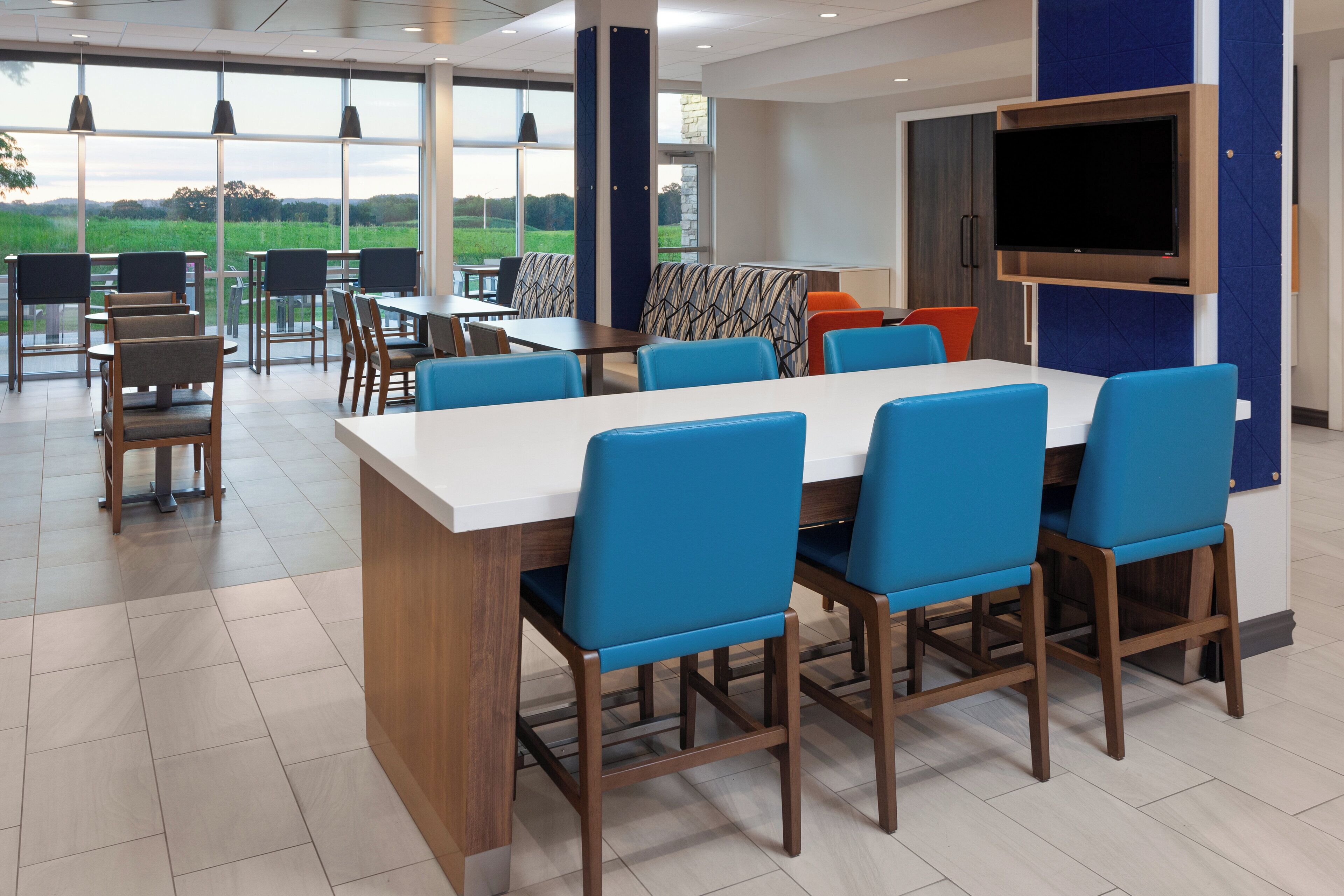 A modern dining area with a large table and blue chairs, large windows with a view of greenery, and a mounted TV on the wall.