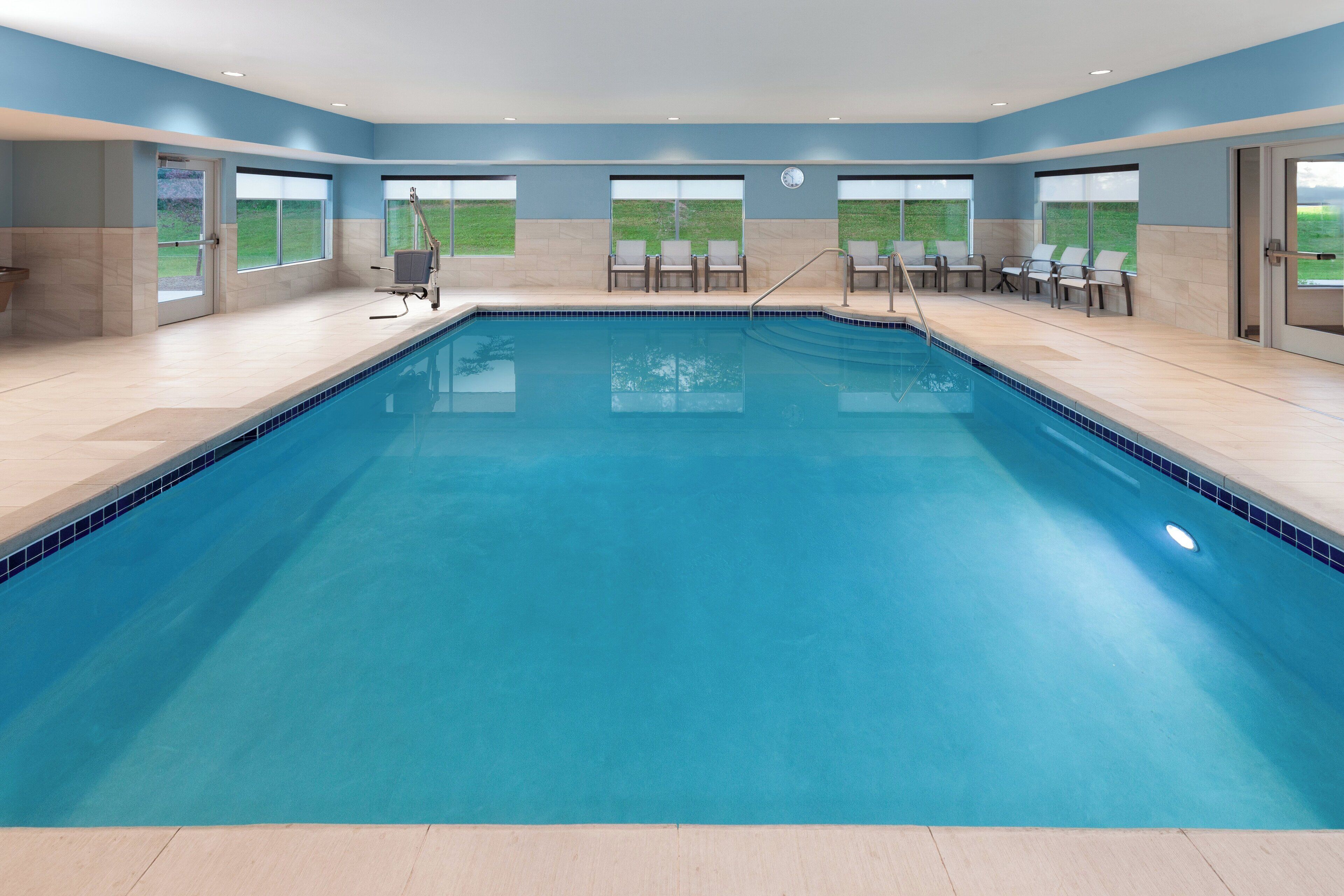 A large indoor swimming pool with clear blue water, surrounded by lounge chairs and a view of a grassy field through large windows.