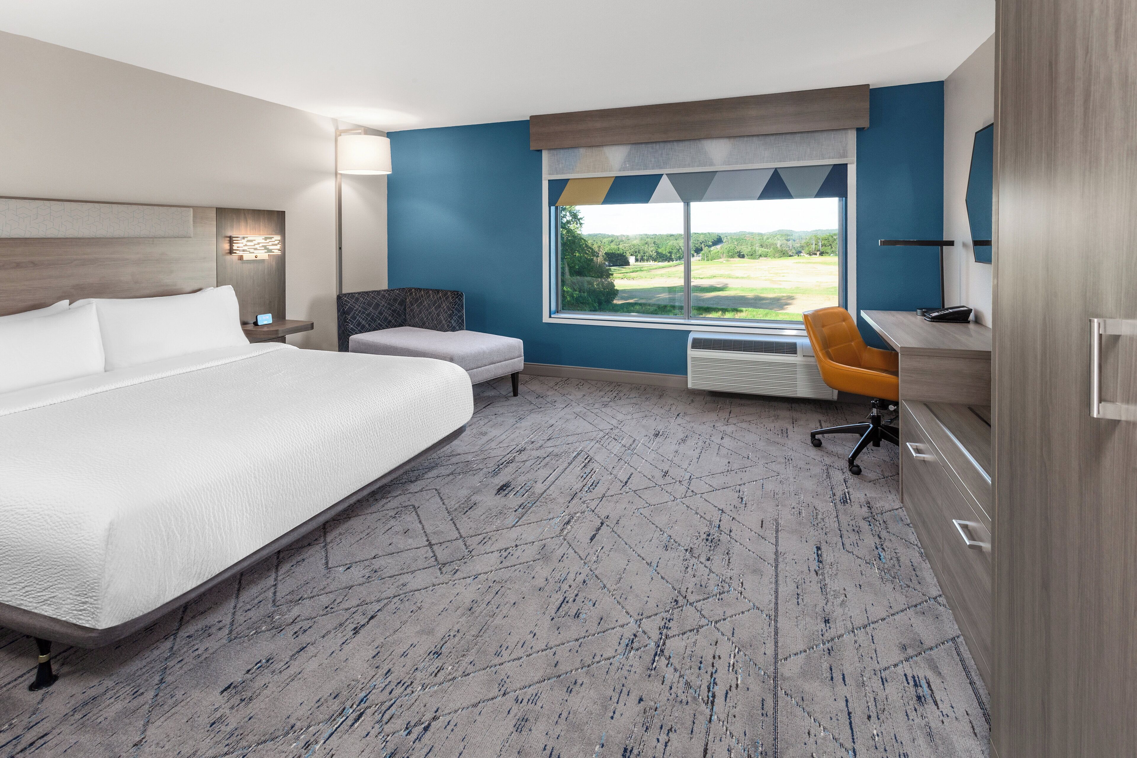 Et moderne hotelværelse med en stor seng, et skrivebord med stol, et vindue med udsigt til grønne områder og en fast installeret varmeovn.