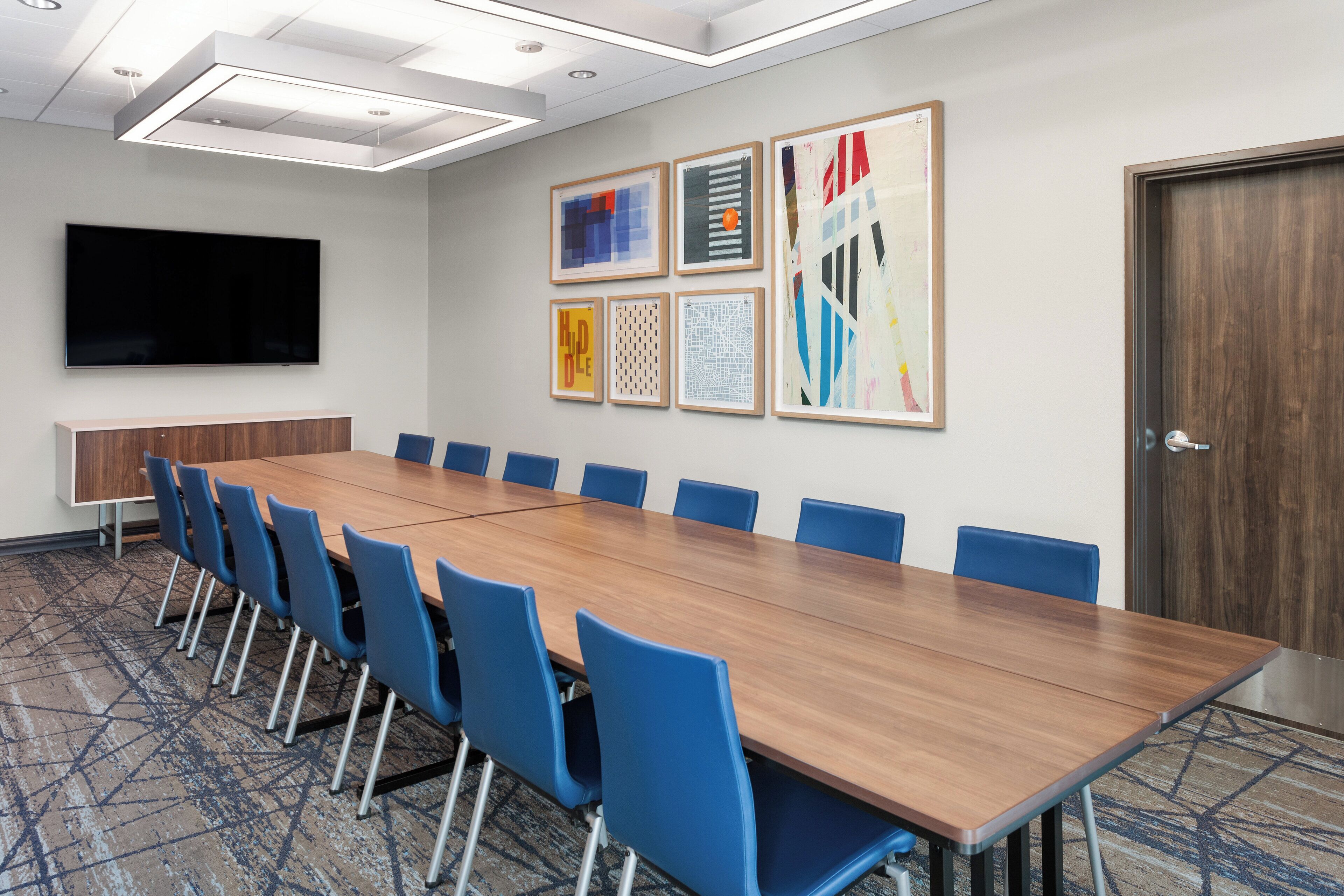 Une salle de conférence avec une longue table en bois, des chaises bleues, un téléviseur à écran plat et des œuvres d’art encadrées accrochées au mur.