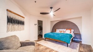 Ein Schlafzimmer mit einem Bett, einem Deckenventilator, Holzboden und einem Wandbehang mit Muster.