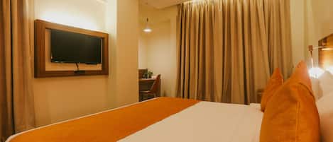 A hotel room with a large bed, a flat-screen TV mounted on the wall, a desk with a chair, and a window with curtains.
