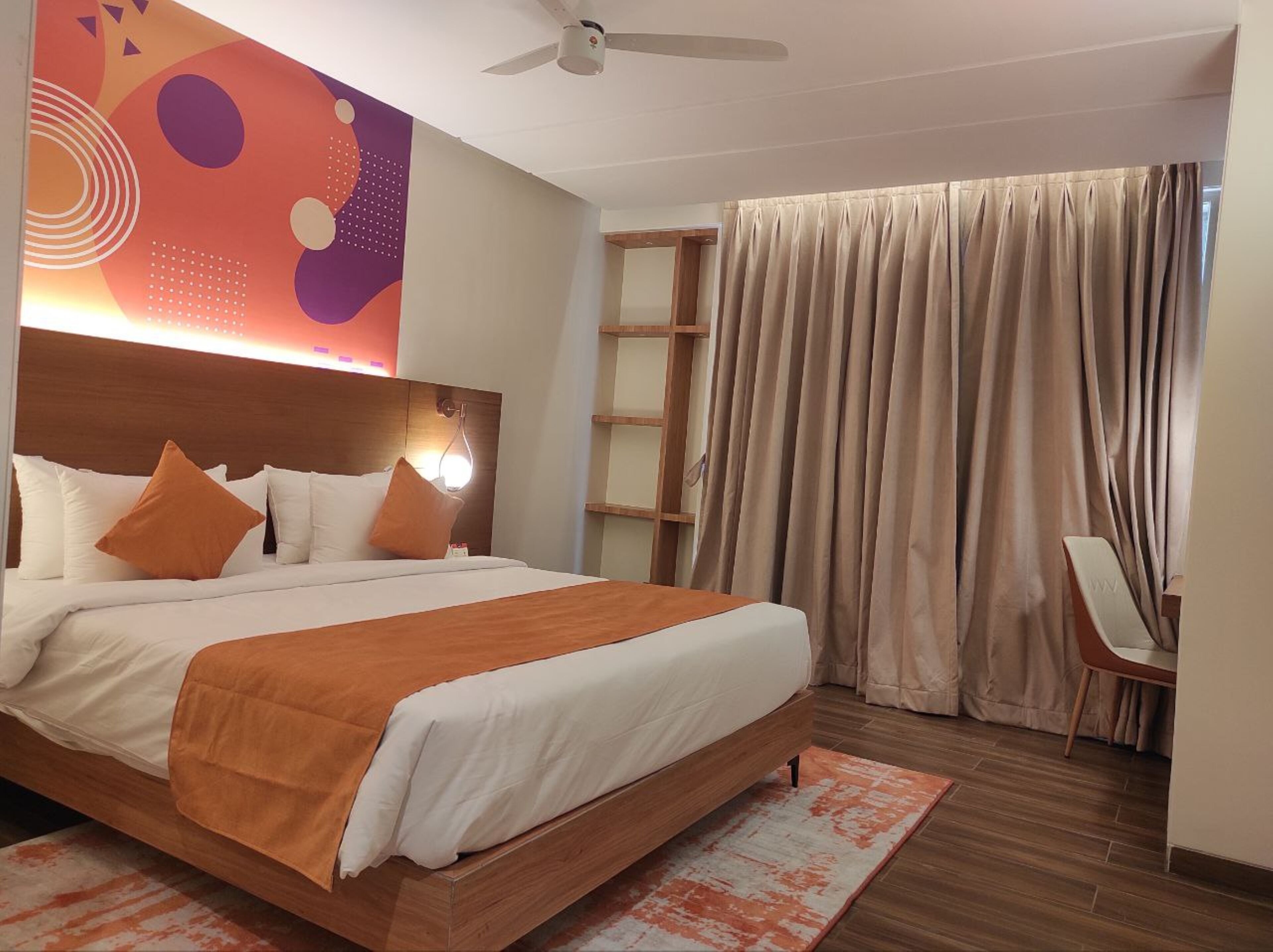Ein modernes Hotelzimmer mit einem großen Bett, einem Deckenventilator, einem Wandbild und Blick ins Freie durch Vorhänge.