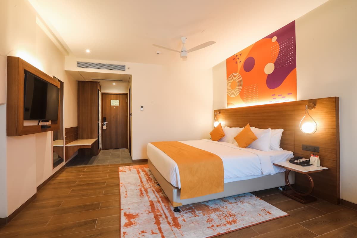 A modern hotel room with a large bed, a bedside table with a lamp, a TV mounted on the wall, and a ceiling fan.