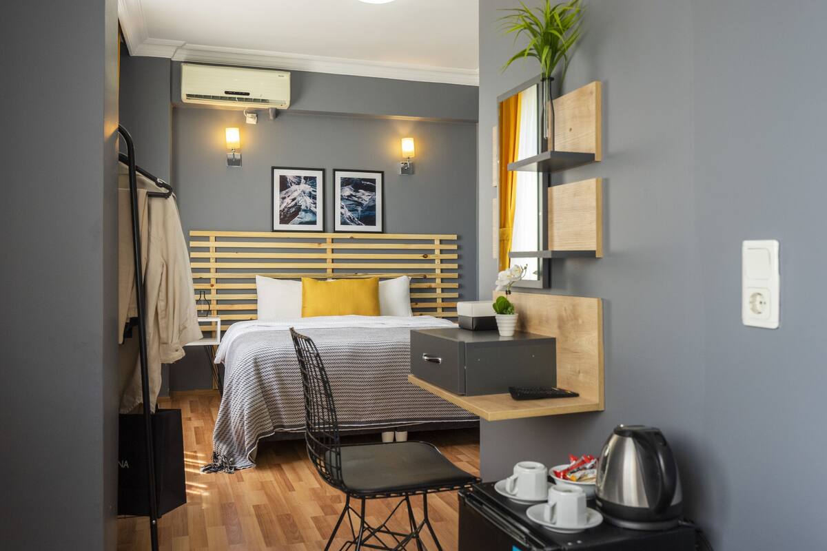 Une chambre d’hôtel moderne équipée d’un lit, d’un bureau, d’une chaise, d’une petite table avec une bouilloire et d’une étagère avec une plante.