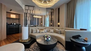A modern hotel room with a large sectional sofa, a round coffee table with flowers, a small side table, and a kitchen area in the background. - LYFE Hotels Bhubaneswar  (Bhubaneshwar)