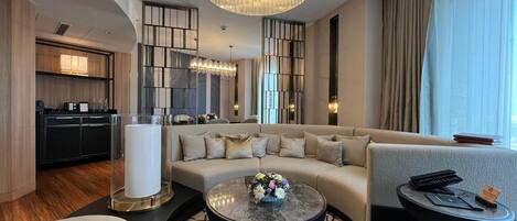 Een moderne hotelkamer met een ruime hoekbank, een rond bijzettafeltje met bloemen, een klein nachtkastje en een keuken in de achtergrond.