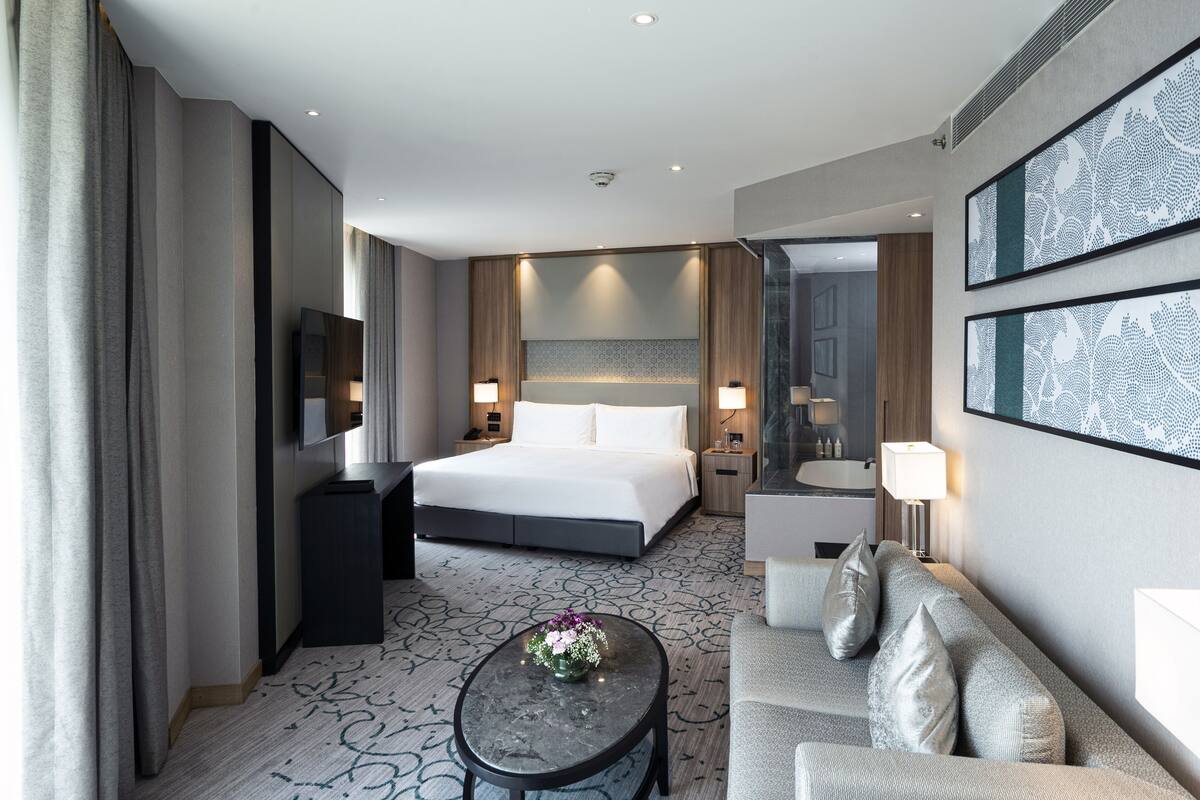 Ein modernes Hotelzimmer mit einem großen Bett, einer Sitzecke inklusive Sofa und Couchtisch, einem Fernseher und einem Badezimmer mit Spiegel und Waschbecken.