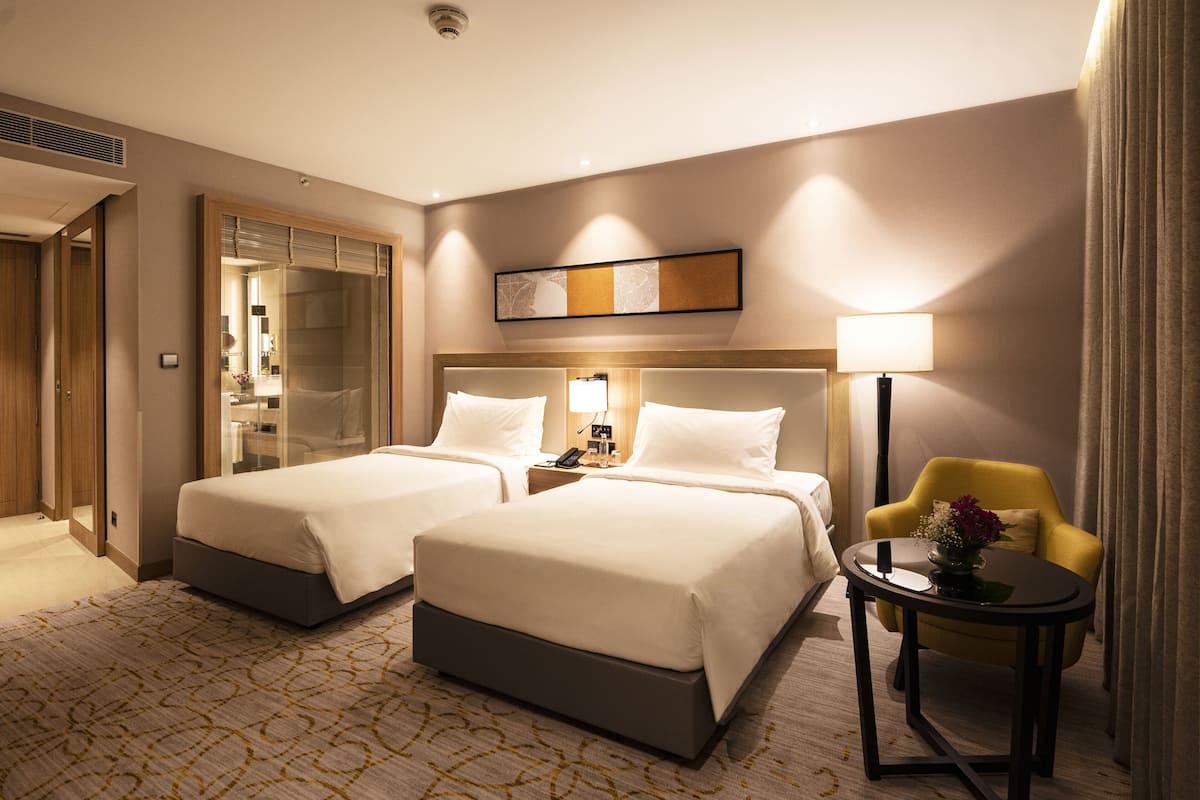 Ein Hotelzimmer mit zwei Betten, einem kleinen Tisch mit Lampe, einem Sessel und einer Wanddekoration.
